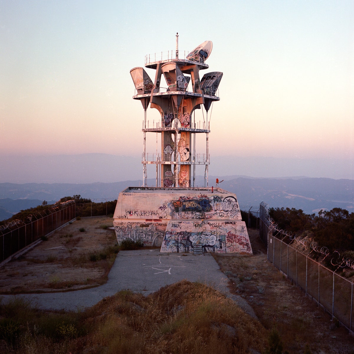 A photo of an abandoned microwave tower with graffiti all over its decaying sides. The tower is surrounded by a barbed wire fence, semiotically acting as both a beacon and a warning to stay away.